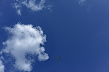 A small propeller airplane flying high in a clear blue sky with scattered white clouds, captured from the ground in broad daylight.