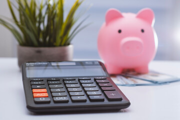 Pink piggy bank wearing glasses on a pile of US hundred-dollar bills with a calculator in the...