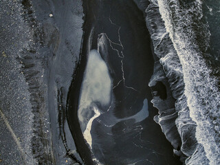 Aerial view of black sands meeting the frothy, white-tipped waves of the ocean, creating a stark contrast with an inlet of milky, pale water, Iceland.