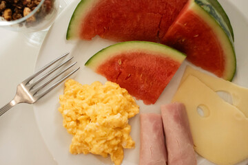 Close up of a Breakfast menu. Scrambled eggs, watermelon, ham and cheese, served on a white plate, with granola on the side, all against a white background. The shiny fork rests on the plate.