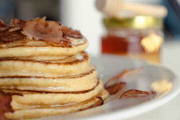 Close-up of a stack of homemade pancakes on a white plate. A small jar of honey in the background, with a golden bee hanging from it. Agave syrup drips from the top. Bacon Pancake day.