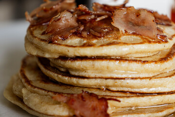 Close-up of a stack of homemade pancakes on a white plate. A small jar of honey in the background, with a golden bee hanging from it. Agave syrup drips from the top. Bacon Pancake day.