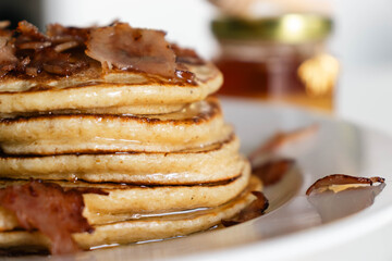 Close-up of a stack of homemade pancakes on a white plate. A small jar of honey in the background, with a golden bee hanging from it. Agave syrup drips from the top. Bacon Pancake day.