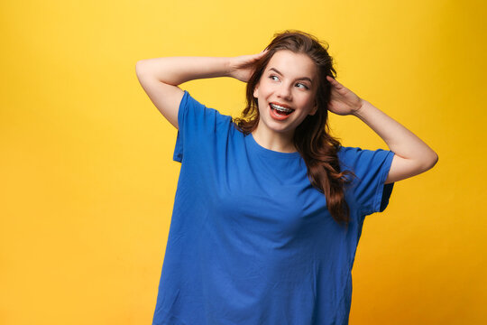 Young girl with dental braces in blue t-shirt, hands behind head, smiling, looking away on yellow backdrop. Dental care services, orthodontic clinics, teen health, lifestyle blogs, youth advertising