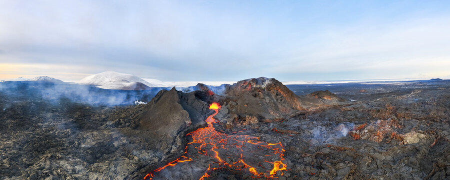 Aerial view of fiery lava flows through jagged, dark volcanic rock towards distant snowy peaks under a pale sky, Grindavik, Grindav&Atilde;&shy;kurb&Atilde;&brvbar;r, Iceland.