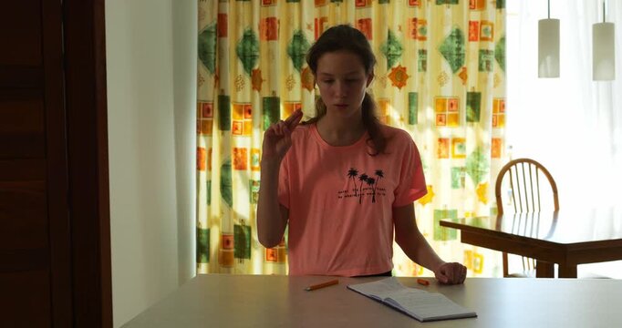 Woman carefully recalls hand movements while studying sign language. Studious brunette interpreter refers to notepad practicing finger gestures near table