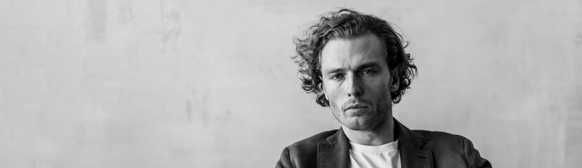 Banner. Young sexy gorgeous handsome fit man in a white shirt sitting on a couch in the studio. Grey background, loft interior, studio. Causal stylish men's clothes. Idea for wedding groom photoshoot