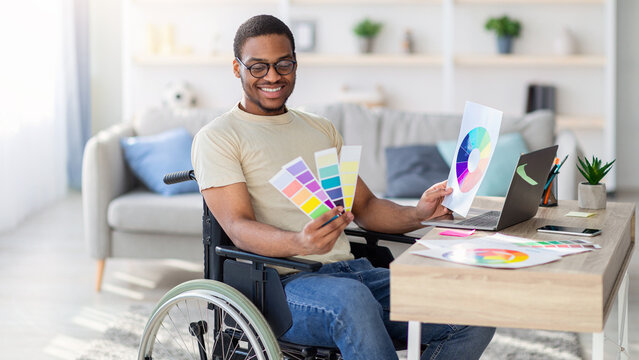 Black guy in wheelchair working as graphic designer online, holding color swatches palette, using laptop at home. Young man with disability having freelance illustrator or developer job