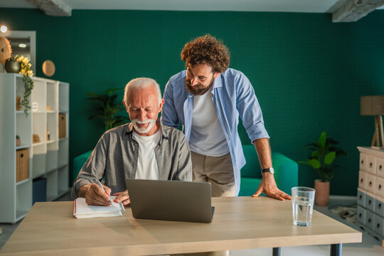 Senior man taking notes while son is helping him with laptop