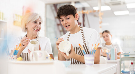 Cheerful elderly woman and young guy, participating in pottery masterclass, sitting together at...