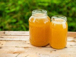 Two glass jars of golden bee honey are on a wooden table, green grass field and sky in the background. Fine organic product concept. Agriculture and food industry.