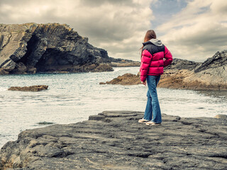 A woman overlooking the ocean and rough stone coast. She is wearing a red jacket and blue jeans....
