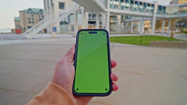 Guy Holds Green Screen Phone Over City Pavement With Buildings And Bystanders Nearby. Image Captures Urban Interaction, Navigation, Or Technology-In-Use On A Regular Day.
