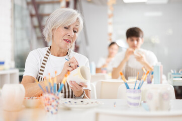 Interested elderly woman attending pottery class, enthusiastically painting on ceramic cup, creating unique custom design. Creative hobby concept