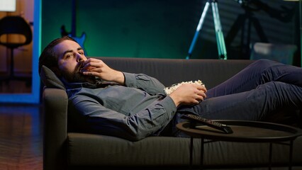 Smiling man lounged on couch eating popcorn while watching movie on streaming service, enjoying...