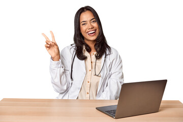 Young Filipino doctor with laptop in studio joyful and carefree showing a peace symbol with fingers.