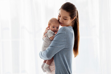 Beautiful young mother holding her adorable newborn baby while standing near window at home, loving...