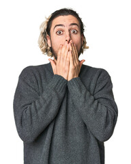 Young Caucasian man in studio setting shocked, covering mouth with hands, anxious to discover...