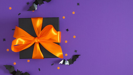 Halloween gift box, black with orange bow, surrounded by bats, pumpkins and on purple background. banner