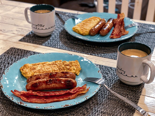 Classic American breakfast with sausages, bacon, folded omelet, and coffee on a rustic table setting.