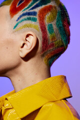 Woman with colorful painted hair and shaved sides wearing a bright yellow textured jacket against a...