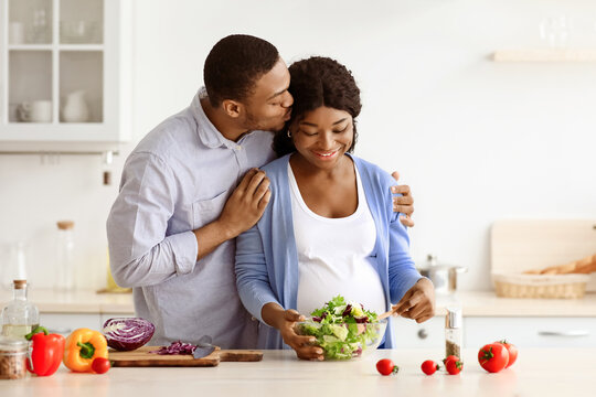Happy african american pregnant couple cooking healthy meal together. Positive expecting black woman and man making dinner at home, making salad from fresh vegetables, kitchen interior - Powered by Adobe