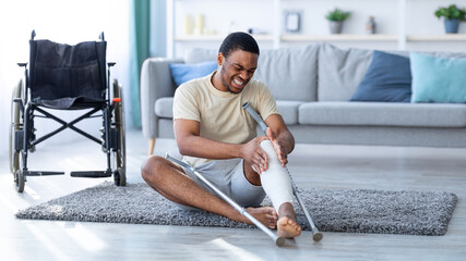 Young black man suffering from pain in plastered broken leg, sitting on floor after falling with...