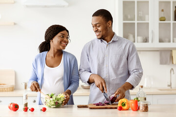 Loving expecting black couple enjoying cooking together, making healthy salad and having conversation, kitchen interior, empty space. Happy african american pregnant wife and husband making dinner