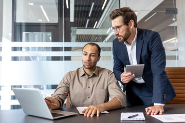 Two business professionals collaborating in an office setting, discussing data on a laptop and tablet.