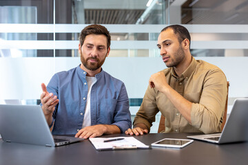 Two men discuss a business strategy, working on laptops at a modern office table.