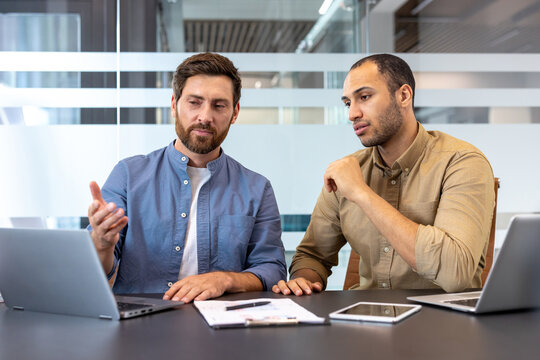 Two men discuss a business strategy, working on laptops at a modern office table.