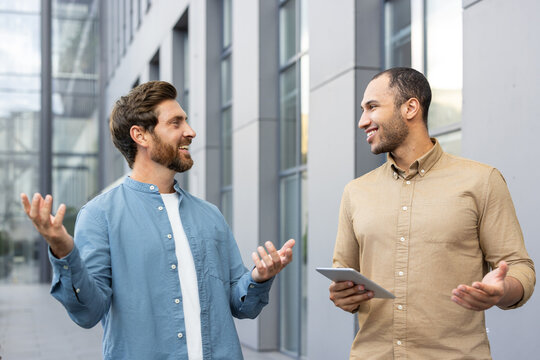 Two diverse businessmen are having a lively conversation outdoors in front of a modern building, possibly discussing a project or deal.