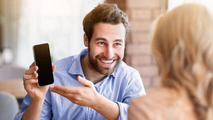 Positive millennial guy showing his girlfriend cellphone with blank screen at coffee shop, mockup...
