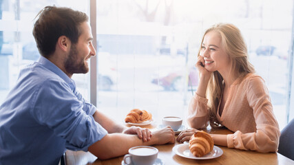 Positive young couple having breakfast together at cafe, sitting near window and enjoying...