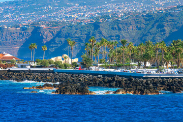 Rocky waves crash along Puerto de la Cruz rugged coast, framed by palm trees lined promenades and distant mountains. Urban charm meets wild Atlantic energy in this vivid Tenerife scene