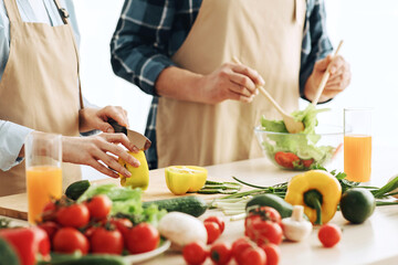 Vegetables for proper nutrition, diet for healthcare. Mature husband and wife in aprons prepare salad in kitchen interior with vegetables and glasses of vitamin juice on table, cropped, close up