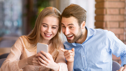 Loving young guy pointing at smartphone during romantic dinner at urban cafe. Cheerful woman and...
