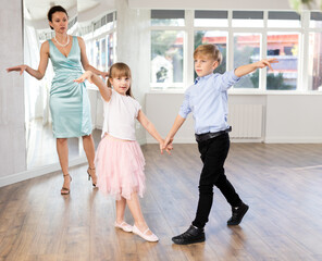 Positive preteen girl and boy practicing samba steps in latin dance studio while female teacher observing and guiding couple..