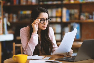 Pensive woman entrepreneur working with papers and laptop computer at cafe, having problem and...