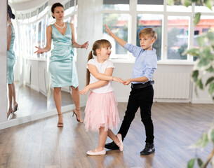 Positive preteen girl and boy practicing samba steps in latin dance studio while female teacher observing and guiding couple..