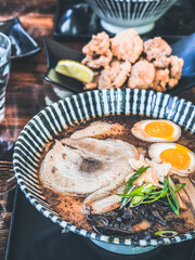 A bowl of Japanese ramen topped with sliced pork, soft-boiled eggs, scallions, and garlic chips, served with karaage fried chicken on the side.