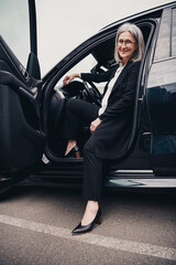 Elegant mature businesswoman sitting in car doorway outdoors, dressed in elegant formalwear, enjoying a productive day.