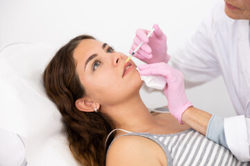 Professional mature female beautician with syringe injecting filler with hyaluronic acid in lips of...
