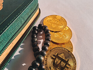 Four golden Bitcoin coins placed beside a green wallet and black prayer beads (tasbih) on white...