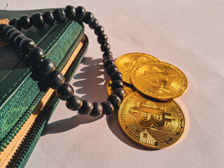 Three golden Bitcoin coins placed beside a green wallet and black prayer beads (tasbih) on white...