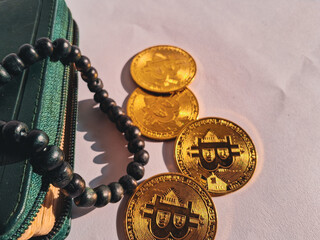 Three golden Bitcoin coins placed beside a green wallet and black prayer beads (tasbih) on white...