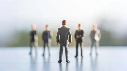 Fototapeta premium A close-up, low-angle view of five business figurines, with the main figure in the foreground facing away from the camera. The team stands together on a reflective surface against a blurred background