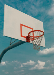 A basketball hoop is shown against a bright blue sky, with the vibrant red rim and net contrasting sharply with the white backboard. 