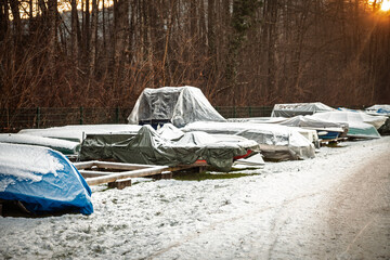Row of small boats covered with tarps and dusted with snow in Austria, stored on land for the cold season beside a forest path. Off season boatyard scene with winter protection and quiet atmosphere.