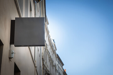 A blank square grey shop sign projects from a building on a French street. The minimalist panel...